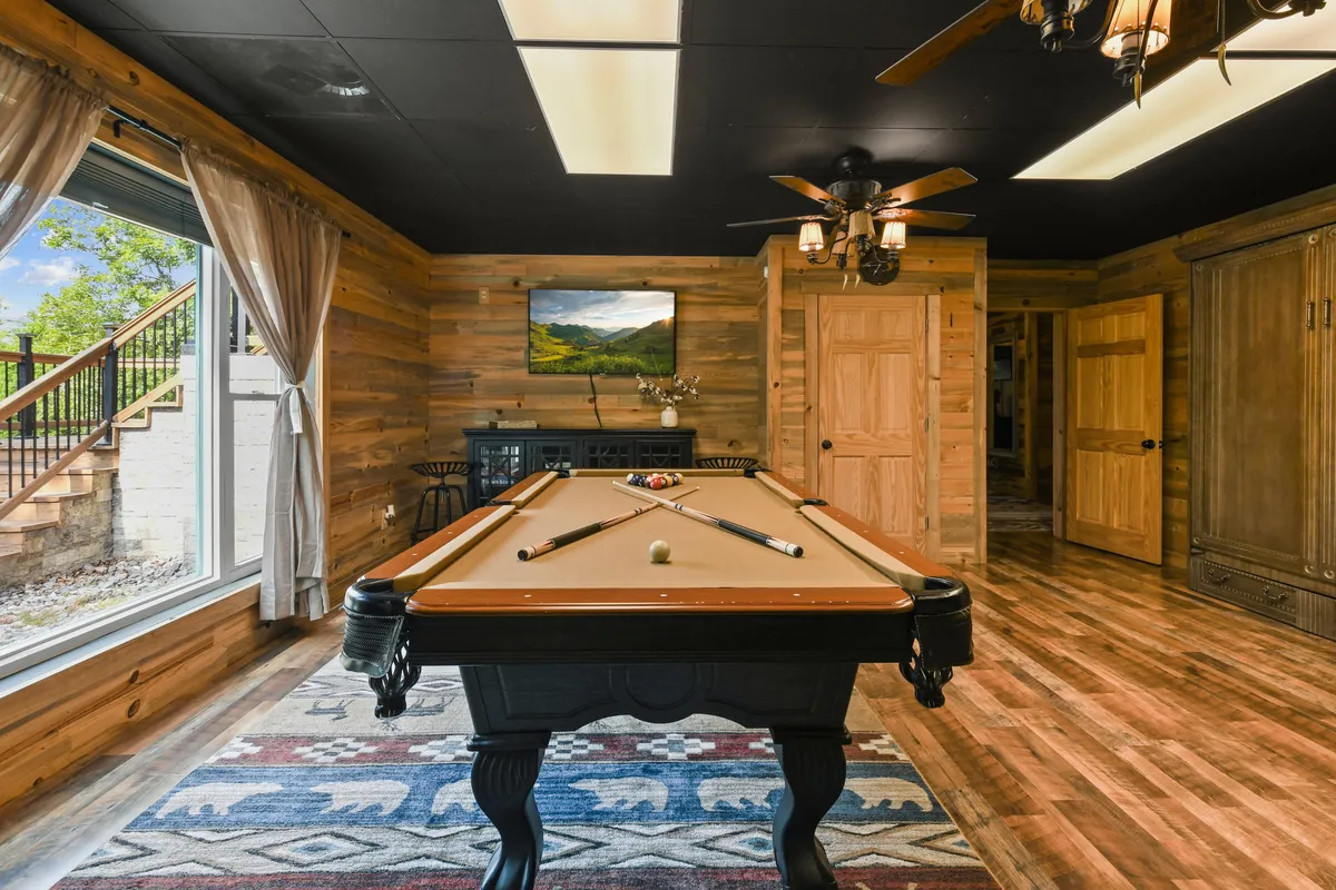 A cozy interior with a pool table as the focal point, set against rustic wooden walls and a large window. The setting invites both friendly competition and relaxed conversation, with natural light enhancing the space.