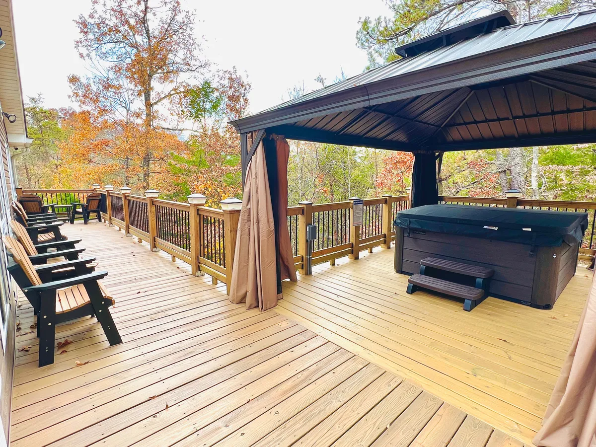 A spacious wooden deck nestled among tall trees, featuring a hot tub beneath a gazebo, comfortable outdoor seating, and glimpses of the surrounding forest. The scene conveys a tranquil atmosphere perfect for relaxation, with the hot tub, deck, and nature seamlessly integrated.