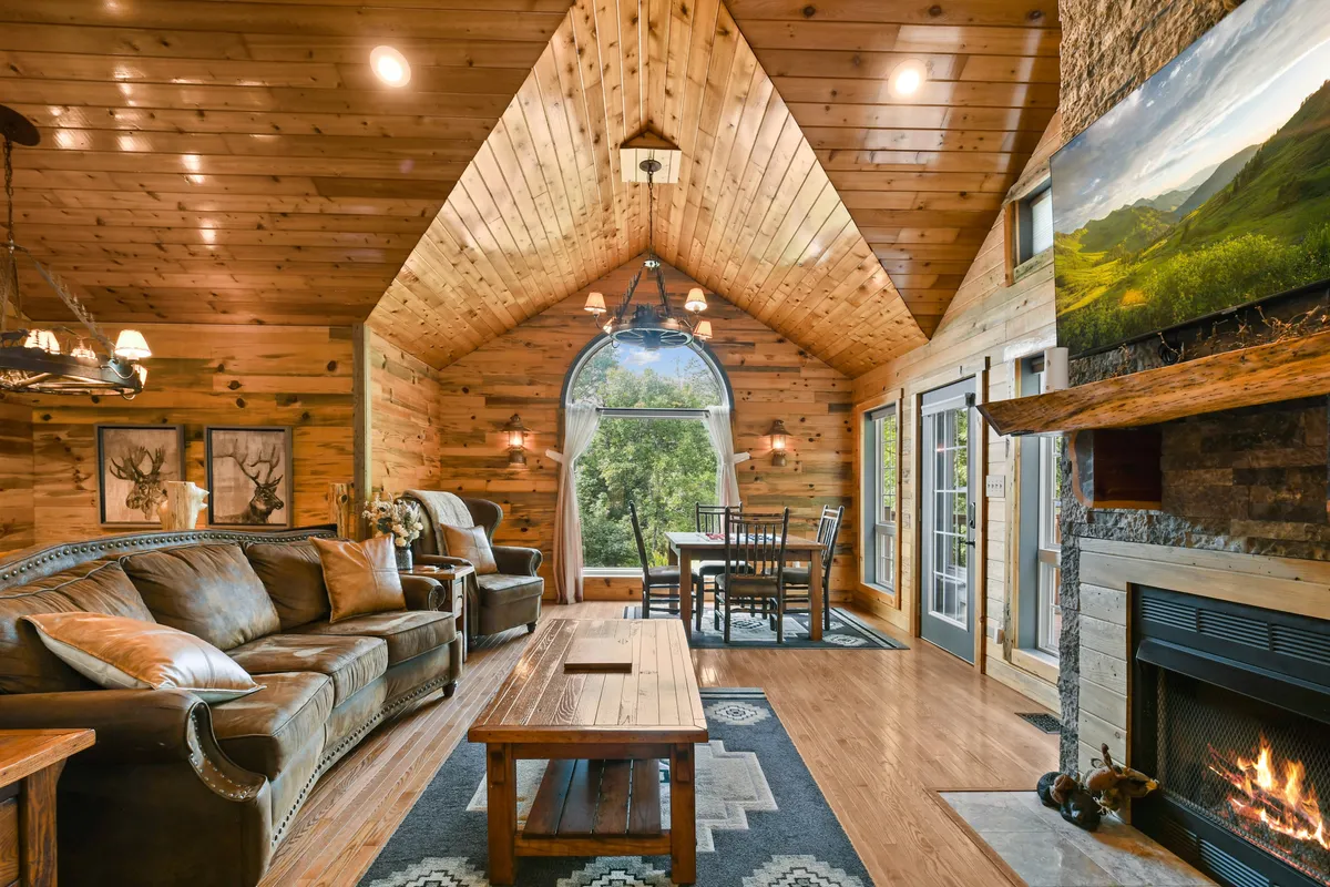 This image presents a sweeping view of the open-concept living and dining area anchored by a striking stone fireplace. Sunlight pours through a large arched window, illuminating a wooden dining table set for a group and a plush leather sofa perfect for family gatherings. The warmth of natural wood accents and the welcoming fireplace define the heart of the space.