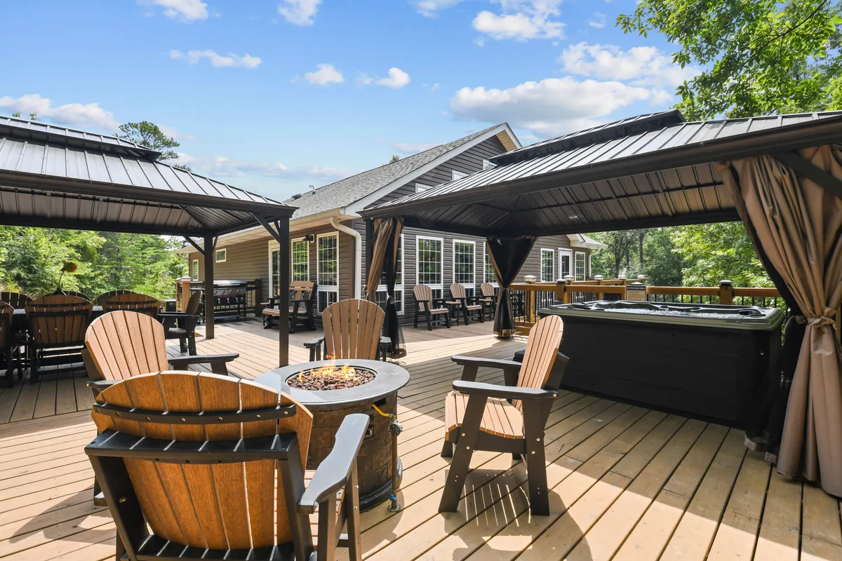 A broad view of the outdoor living space with a large deck, inviting hot tub, several covered gazebos, a fire pit encircled by comfortable chairs, and forested views beyond. The image suggests ample space for gatherings, relaxation, and stargazing.