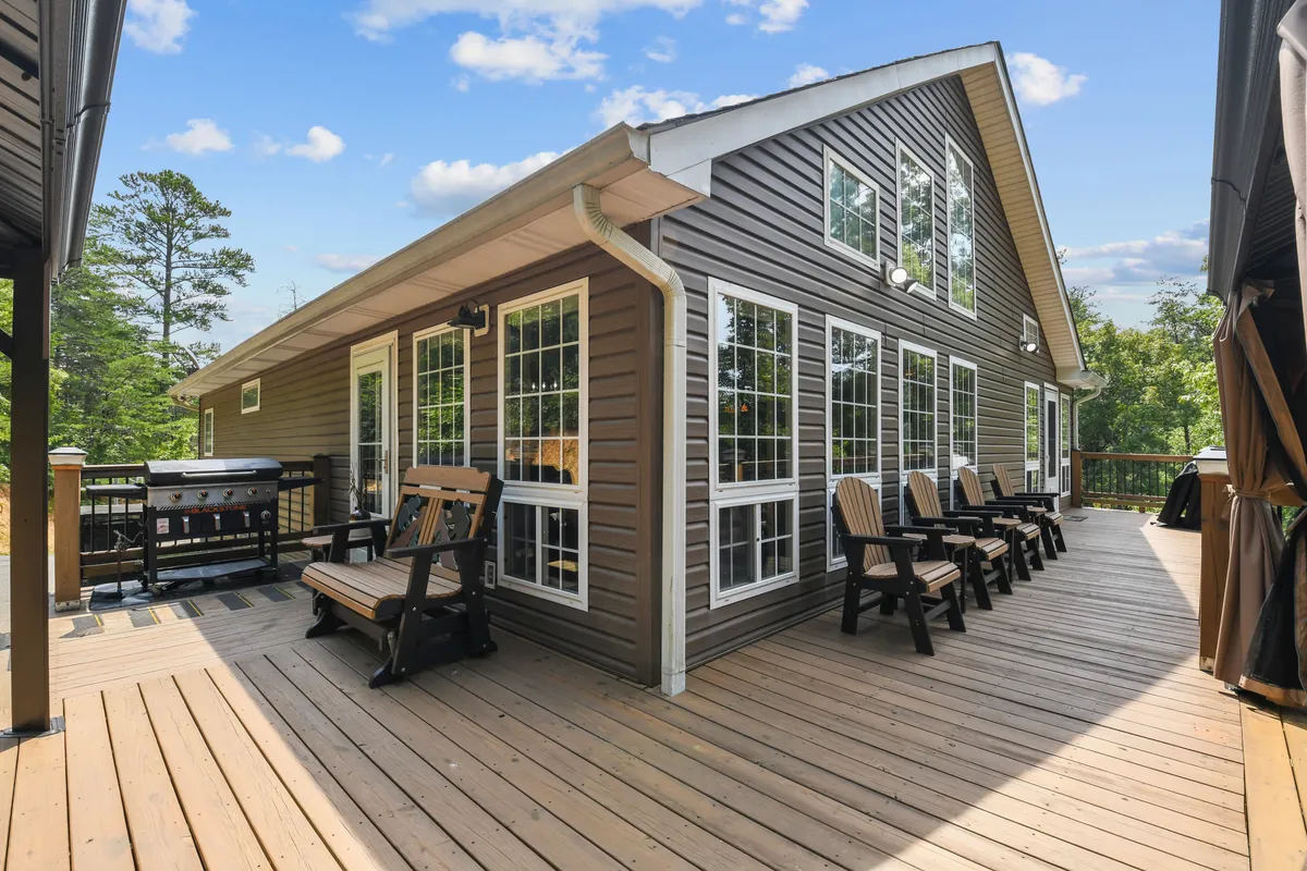 A spacious deck with comfortable outdoor seating, set against a backdrop of windows and lush surrounding trees. The scene hints at open sky and the tranquility of nature, with ample space for relaxing, entertaining, or dining outdoors.