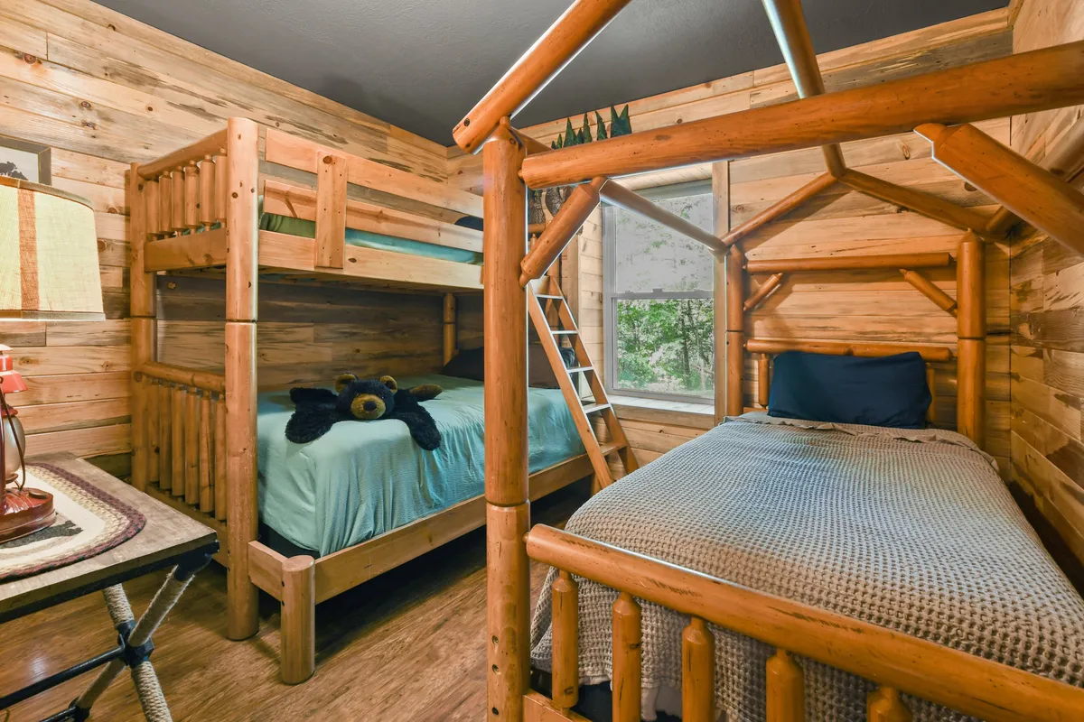 A playful, whimsical bedroom with a twin-over-queen bunk bed, a single bed, and charming wooden furniture. A teddy bear and plenty of natural light add to the room’s inviting, family-friendly atmosphere. The window provides a peek at the outdoors.