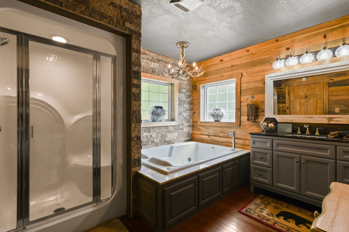 A serene bathroom featuring a large walk-in shower with glass enclosure, a spacious vanity with mirror, and a soaking bathtub. Warm wooden walls and a sparkling chandelier set a calm, refined mood ideal for self-care.