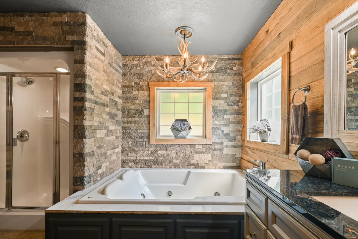A tranquil bathroom retreat with a luxurious jetted jacuzzi tub, elegant chandelier, textured stone wall, and rustic wood accents. A large window frames peaceful nature views, creating a spa-like setting perfect for unwinding.
