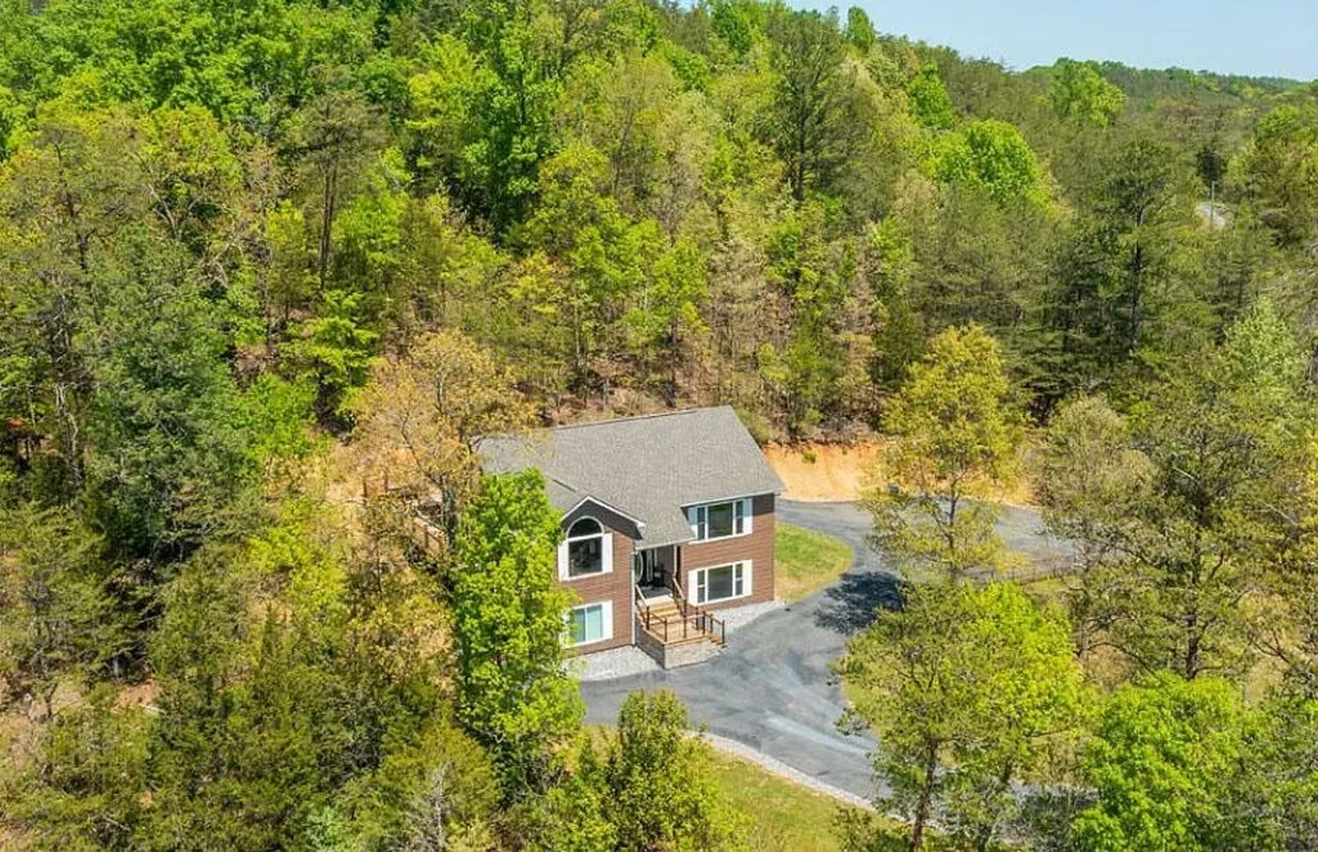 A wide view of the house nestled among tall trees, with an inviting driveway and a wraparound porch. The natural setting emphasizes the connection between the home and its mountain surroundings, suggesting opportunities for hikes and outdoor play.