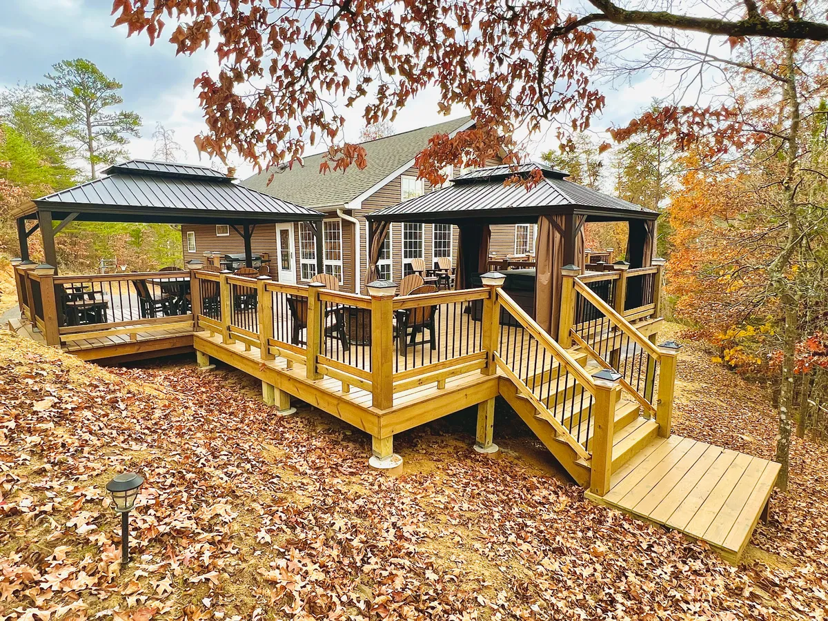 A spacious wooden deck surrounded by trees featuring outdoor furniture and gazebos, perfect for group gatherings and outdoor meals. The setting offers a welcoming, natural environment with ample room for guests to dine, relax, or stargaze together, reflecting a blend of comfort and the outdoors.