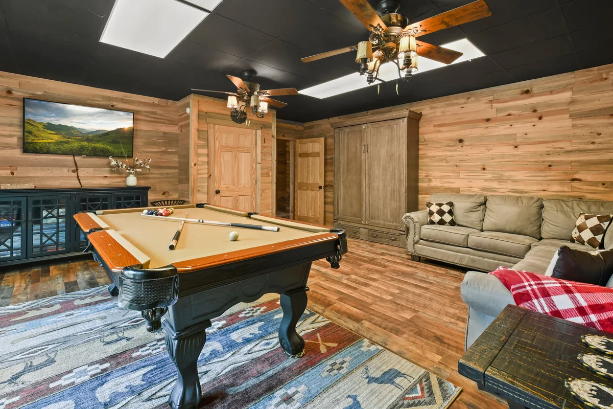 A warmly lit game room featuring a pool table, sofa, television, and wooden walls. The space exudes a relaxed, welcoming vibe and offers ample area for playing games, lounging, and socializing.