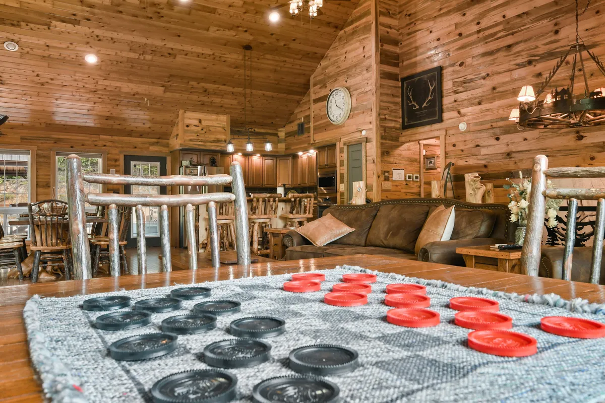 A game-focused room with a checkerboard game on the table, rustic wooden furniture, soft lighting, and a large wall clock. The layout encourages board games, cozy gatherings, and downtime in a playful yet relaxed environment.