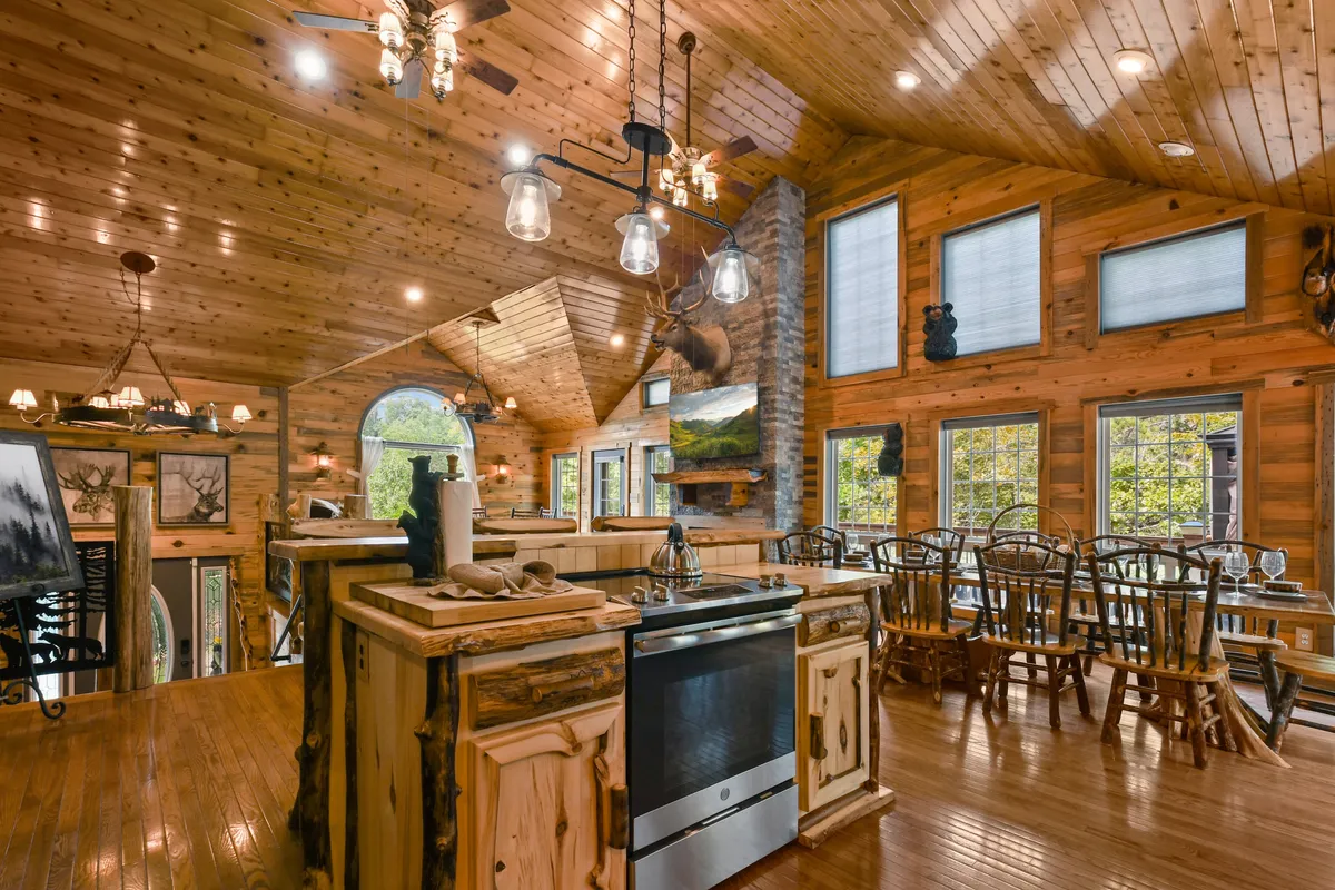 A wide view of the kitchen and dining area features a prominent kitchen island, a grand wooden table, a stone fireplace, and exposed wooden beams. Large windows offer woodland views, and the layout encourages both culinary creativity and communal dining.
