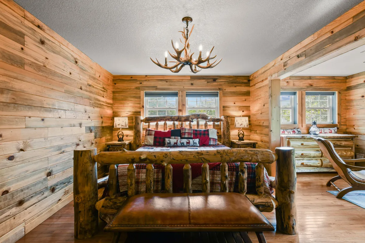 A warm, inviting bedroom featuring a rustic log bed, soft lighting from a chandelier, and wooden walls. Sunlight filters in through the windows, casting a tranquil glow across the space. The room exudes coziness and comfort, ideal for restful sleep after a day of adventure.