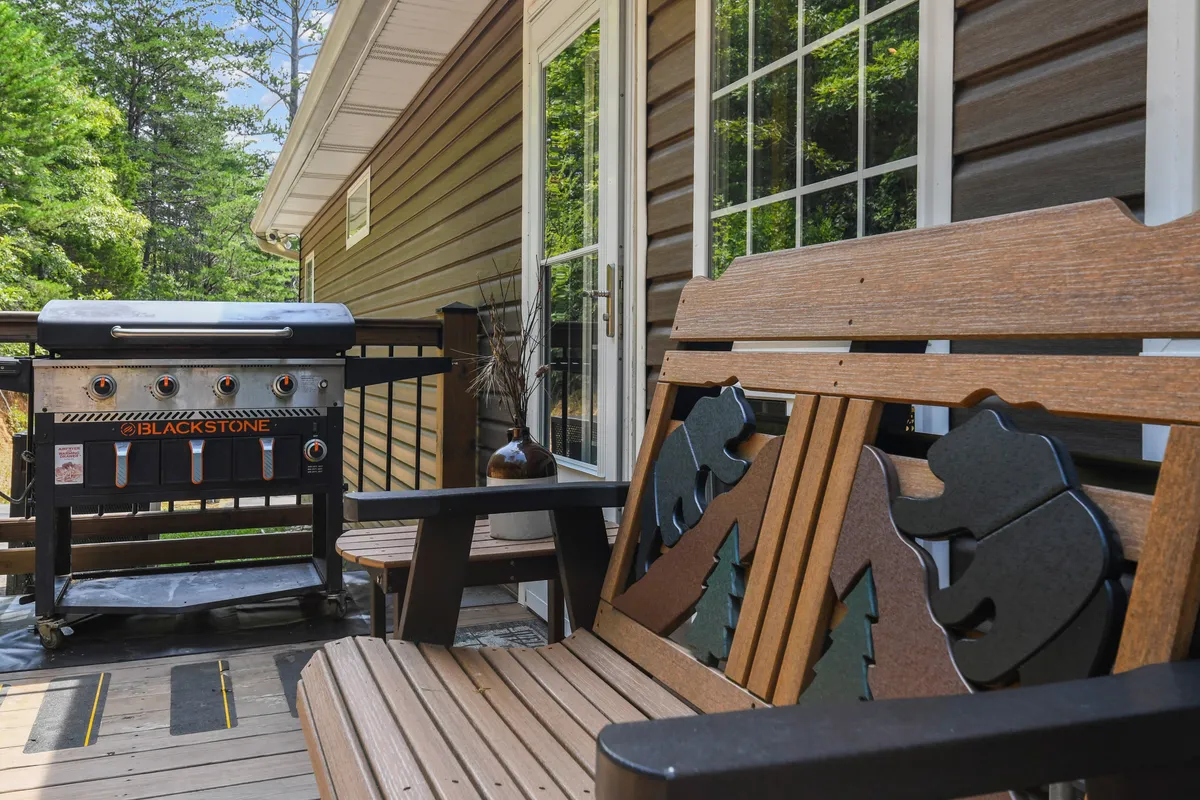 A cozy exterior scene showing a barbecue grill and a rustic wooden bench with bear carvings, set against a backdrop of greenery. This inviting setup highlights a spot for grilling, relaxing, and sharing conversations in a scenic outdoor space.